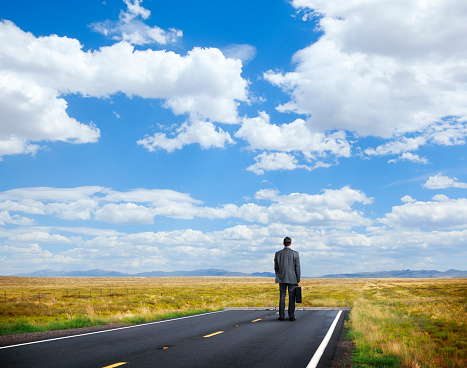 A businessman stands at the end of a road as he looks out towards a beautiful panorama of grassland and a cloud filled sky.  The road has come to an abrupt end as he stands holding his briefcase wondering where he is to go from here.