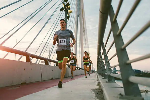 Runners running marathon in the city. They are running over the bridge at sunset. Wearing numbers on their sport clothes.