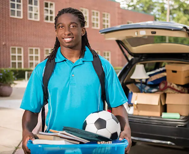 African descent boy heads off to college.  The 18-year-old is unpacking his car as he moves into the college campus dorm.  He is excited to start his school adventures. He carries a backpack and a tub full of his belongings.  Life events.  Back to school.