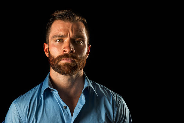 One 25 year old, bearded male wearing a casual button up shirt shot against a black background in studio having the expression of interest on his face.