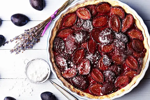 Homemade pie with plums, lavender and sieve with sugar powder on the table with white and blue wooden background. Baked and raw plums. Top view