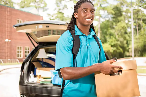 African descent boy heads off to college.  The 18-year-old is unpacking his car as he moves into the college campus dorm.  He is excited to start his school adventures. He carries a backpack and moving box.  Back to school.