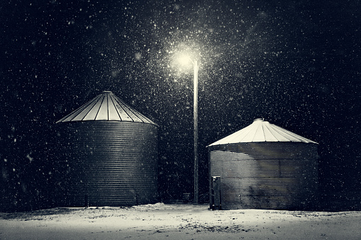 Grain storage bins in a night time winter snow storm on the farm. Black and white photograph with warm tones of white. Snow covers the ground, and light from a single light pole shines through the blizzard. No people in this high resolution photo withi copy space and horizontal composition.