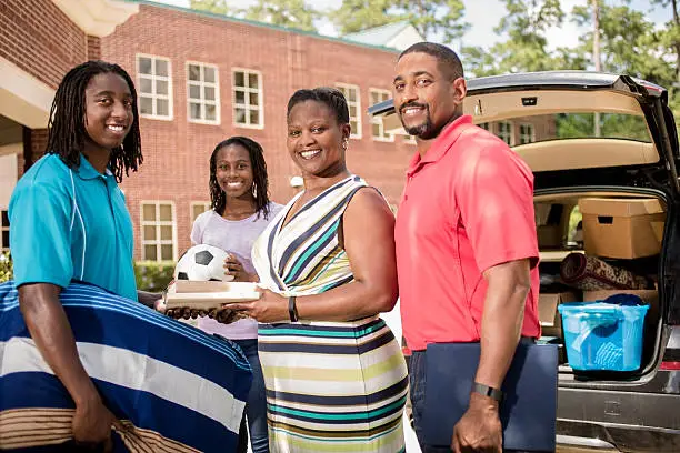 African descent boy heads off to college.  The 18-year-olds' parents and sister are all helping him unpack his car as he moves into the college campus dorm.  He is excited to start his school adventures. He  carries a pillow and textbooks.  Family events.  Back to school.