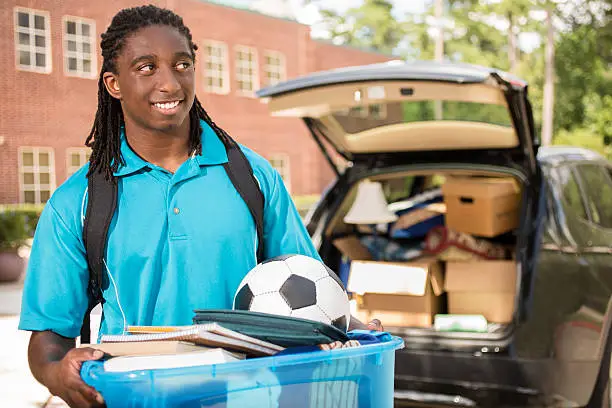 African descent boy heads off to college.  The 18-year-old is unpacking his car as he moves into the college campus dorm.  He is excited to start his school adventures. He carries a backpack and a tub full of his belongings.  Life events.  Back to school.
