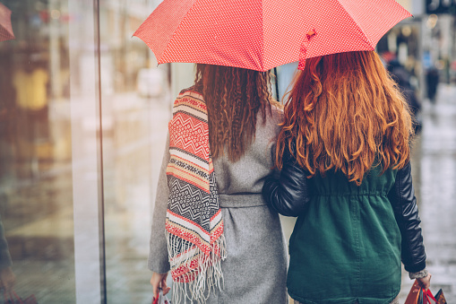 Friends walking on city street while raining. Holding red umbrella and shopping bags. Wearing warm clothing.