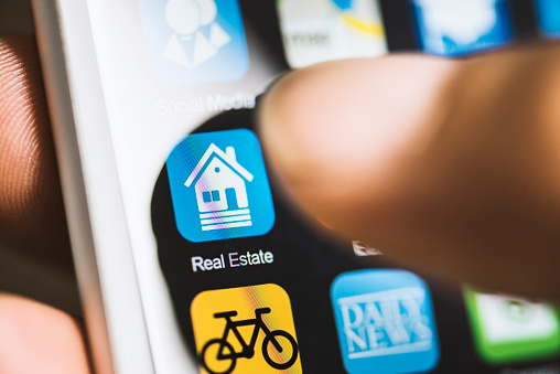 Hand holding phone, touching real estate application icon with thumb