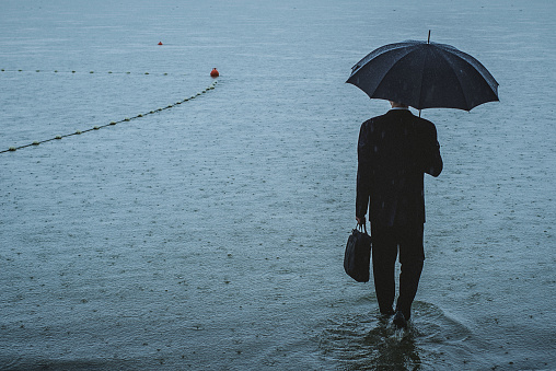 Rear view on a handsome man wearing suit and holding umbrella and a briefcase, standing on the waterfront on a rainy day. The man waded ankle-deep in water. Selective focus, three quarter length, copy space has been left.