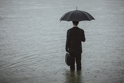 Rear view on a handsome man wearing suit and holding umbrella and a briefcase, standing in the water on a rainy day. The man waded knee-deep in water. Selective focus, three quarter length, copy space has been left.