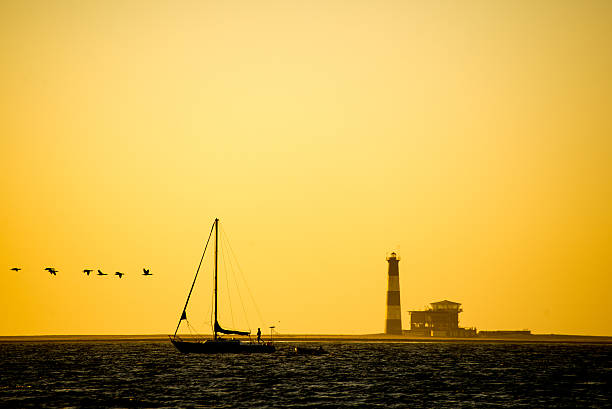 Sailing Yacht at Sunset A sailing yacht sails past Pelican Point Lighthouse at Walvis Bay while seabirds also fly  by. sailboat mast stock pictures, royalty-free photos & images
