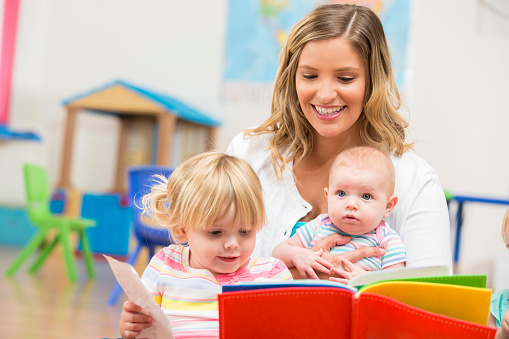 Caucasian, blonde woman wearing a white cardigan is sitting on the floor of a daycare while reading a book to two young girls. She is smiling and holding an infant baby and a toddler. There are toys in the background.