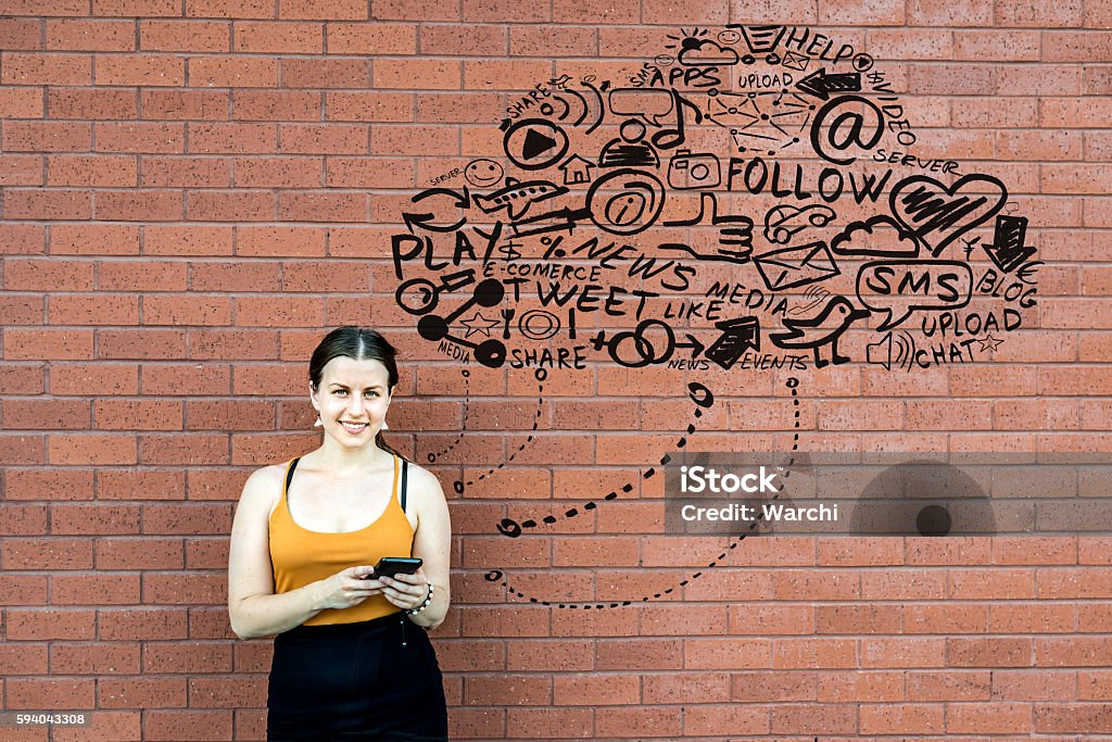 She is connected to the cloud young woman in front of a brick wall using her phone. She is connected to the cloud. The cloud is hand drawn out of internet symbols and icons Brick Stock Photo