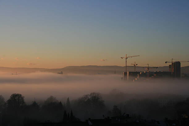 The Old Town of Oslo in the foreground and the new Barcode buildings under construction. A late summer evening sunset and the fog rolling in from the Oslo Fjord. Trees tops of the Old Town graveyard in the foreground. Blue sky and small clouds floating in the orange sunset.