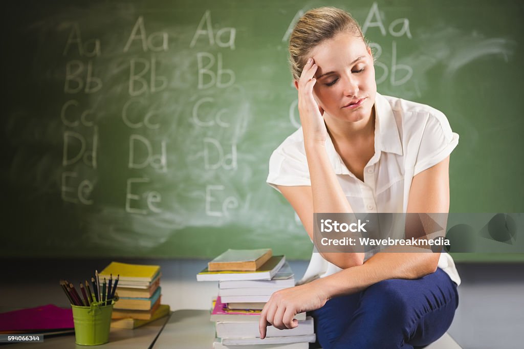 Tensed school teacher sitting in classroom Tensed school teacher sitting with hand on forehead in classroom at school Teacher Stock Photo