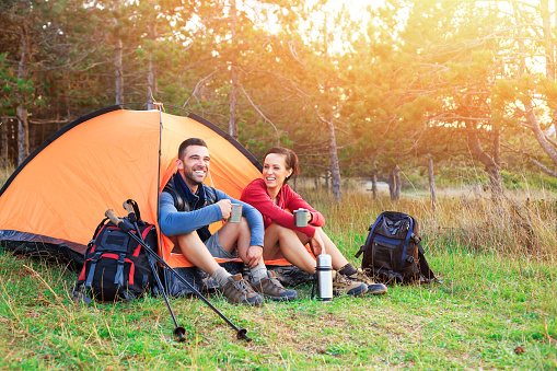 Young couple sitting in orange tent and drinking coffee. Both with backpacks and sweatshirts, hiking shoes, holding a metal mugs. Thermos next to them. A tent, trees and sunlight on background.
