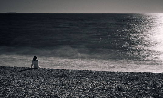 Unrecognizable woman sits on the pebble beach in Marina di Pisa, Italy. Long exposure in the infrared spectrum