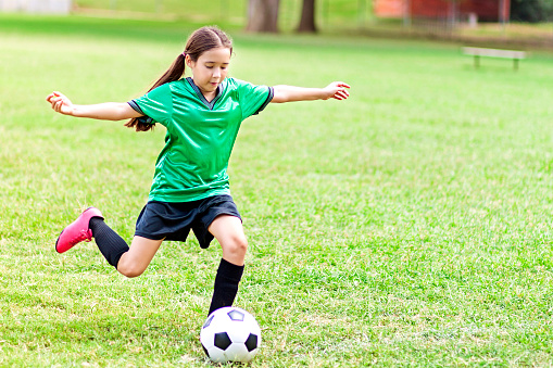 Focused Hispanic girl prepares to kick soccer ball. Her arms are outstetched as she pulls her foot back to prepare to kick the ball. The has long brown hair in a ponytail. She is wearing a green jersey, black shorts, black socks and pink cleats. She is the only player seen in the photo.