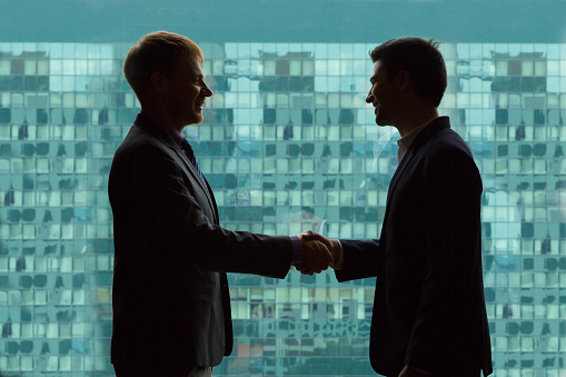 Silhouettes of businessmen shaking hands against big window in modern office building