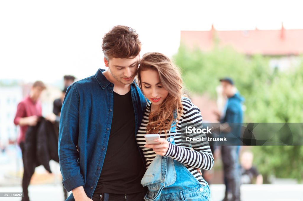 Young couple using smart phone together outdoor Young couple using mobile together outdoor. Group of people in the background. 20-29 Years Stock Photo