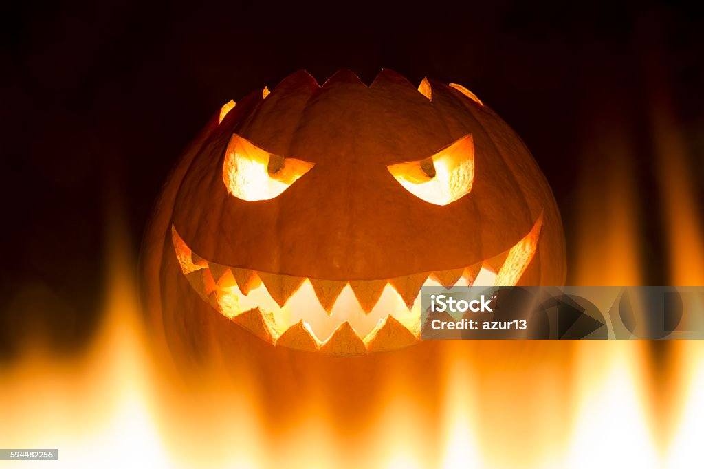 Spooky carved halloween pumpkin in hot burning hell fire flames The big helloween pumpkin has a mad face with glowing eyes and also a glow in its mouth and teeth. Perspective from bottom up. 30-34 Years Stock Photo