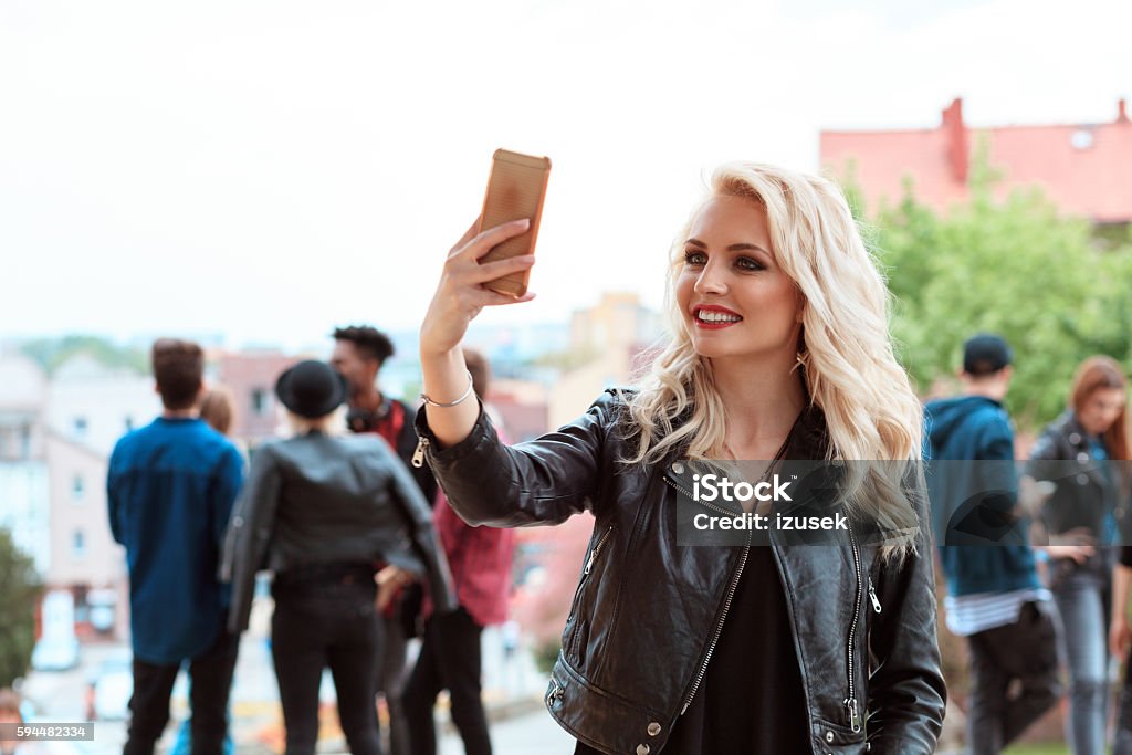 Young woman taking selfie outdoor Young beautiful blonde woman taking selfie outdoor, using smart phone. Multi ethnic group of people in the background. 20-29 Years Stock Photo
