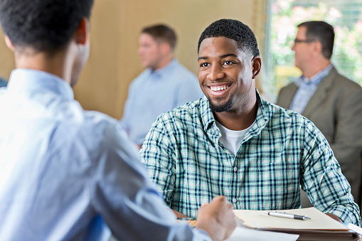 Smiling African American young adult at a job interview in a business center. He is sitting at a desk across from a manager being interviewed for a job. Other applicants are waiting in the background.