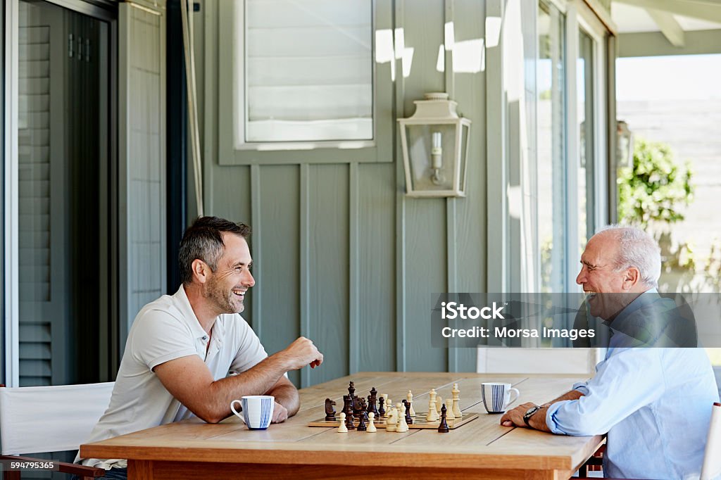 Smiling senior man playing chess with son Side view of smiling senior man playing chess with son on patio Chess Stock Photo