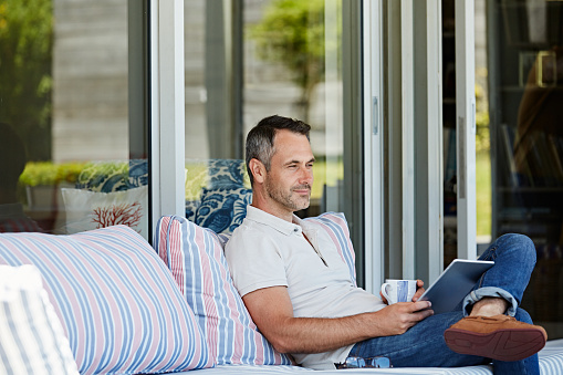 Mature man with digital tablet and coffee cup sitting on sofa at patio