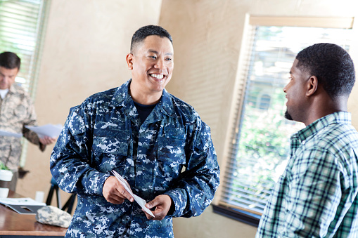 Mature adult Asian male soldier is smiling while working at military recruitment event. He is talking to young adult about joining the armed forces.