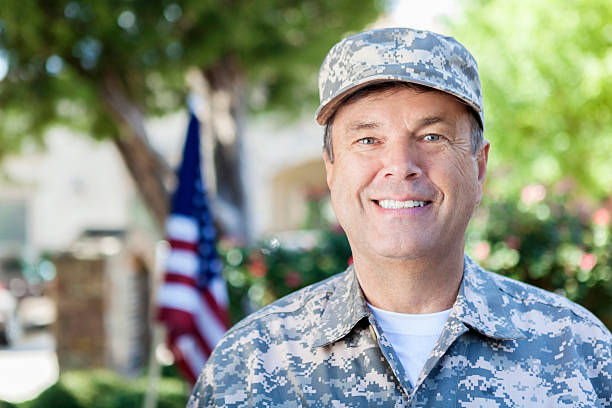 Mature adult soldier in uniform smiling outdoors Mature adult Caucasian man wearing military fatigues including camouflage hat and uniform is smiling and looking at the camera. He is standing outdoors on a sunny day. An American flag can be seen in background. patriotic man stock pictures, royalty-free photos & images