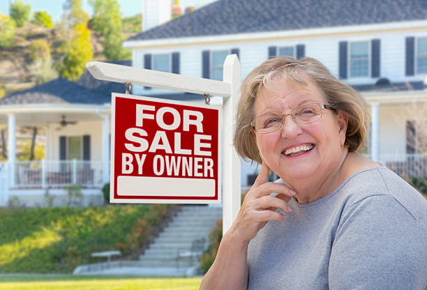 Senior Adult Woman in Front of Real Estate Sign, House Senior Adult Woman in Front of Home For Sale By Owner Real Estate Sign and Beautiful House. house for sale by owner stock pictures, royalty-free photos & images