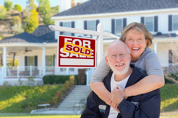Senior Adult Couple in Front of Real Estate Sign, House Senior Adult Couple in Front of Sold Home For Sale Real Estate Sign and Beautiful House. seniors-moving-house stock pictures, royalty-free photos & images