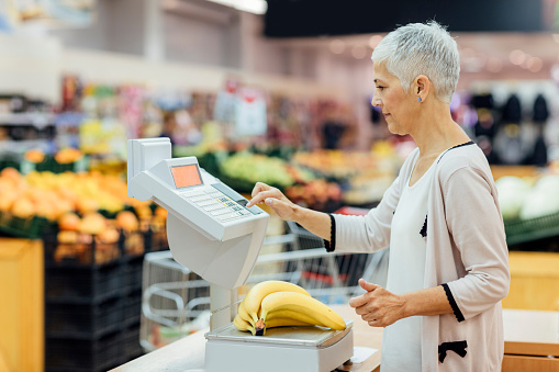 Mature smiling woman shopping in local supermarket. She is shopping groceries, weighing fruits, bananas.