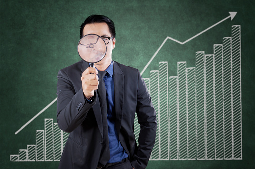 Young businessman looking through a magnifying glass with financial graph background