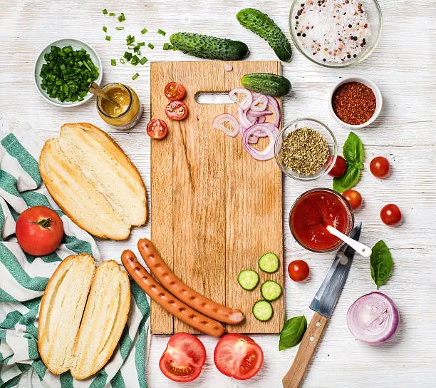 Ingredients for making hot-dogs. Sausages, fresh vegetables, ketchup, mustard, bread and spices with chopping board in center over white painted wooden background, top view, copy space