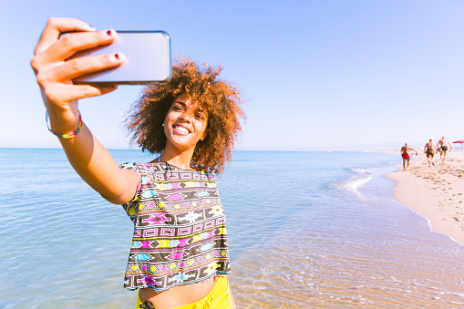 Young black woman taking a selfie at beach.  Twenty years old girl, mixed race caucasian and african black with curly hair. Travel and vacations concepts. Color filter added.