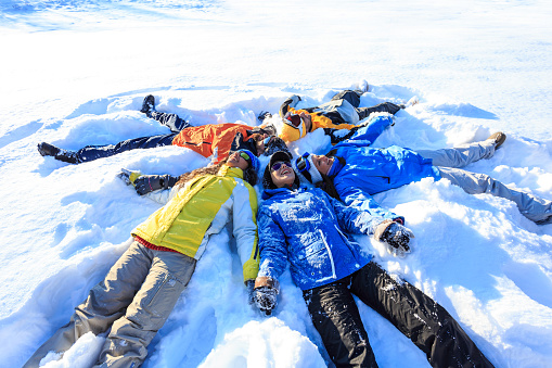 Cheerful young people lying down and making snow angels in the mountain, holding hands. Wears warm clothes and gloves, knit hats and sunglasses. Mountain peak, trees and clear sky on background.