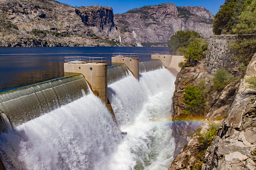 color horizontal photo of O'Shaughnessy (Hetch Hetchy) Dam in Yosemite National Park, California, USA. Water gushing forth from the dam with slight rainbow in foreground, cobalt blue water in mid ground and cliffs and waterfall of Hetch Hetchy Valley in background.
