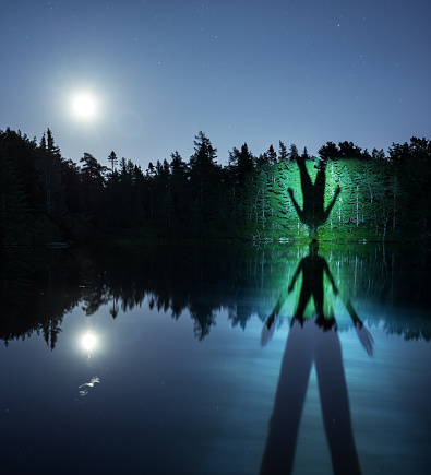 A person's shadow is projected onto the distant shoreline of a lake and reflected in the still surface under the rising Moon.  Long exposure.