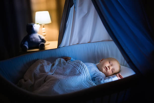 Baby boy sleeping at night Adorable baby sleeping in blue bassinet with canopy at night. Little boy in pajamas taking a nap in dark room with crib, lamp and toy bear. Bed time for kids. Bedroom and nursery interior. crib stock pictures, royalty-free photos & images
