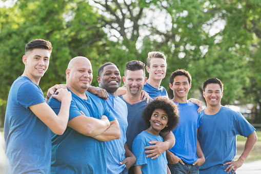 A multi-ethnic group of eight boys and men wearing blue shirts and jeans. They are fathers and son working on a community service project together. The men are in their 40s. The youngest son is 12 years old, the mixed race African American boy standing in the middle looking over his shoulder. The other sons are 17-18 years old.