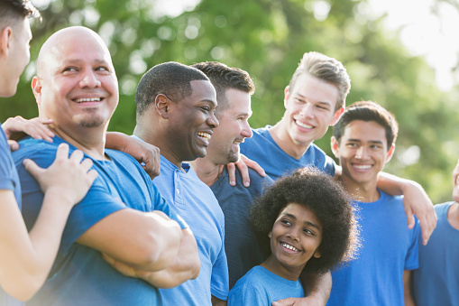 A multi-ethnic group of six people, fathers and their sons. The dads are in their 40s. The main focus is on a 12 year old mixed race African American boy and his dad who are looking at each other.