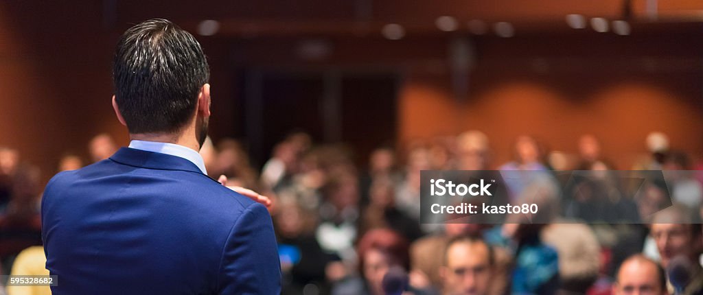 Public speaker giving talk at Business Event. Rear view of speaker giving a talk on corporate Business Conference. Audience at the conference hall. Business and Entrepreneurship event. Panoramic composition. Public Speaker Stock Photo