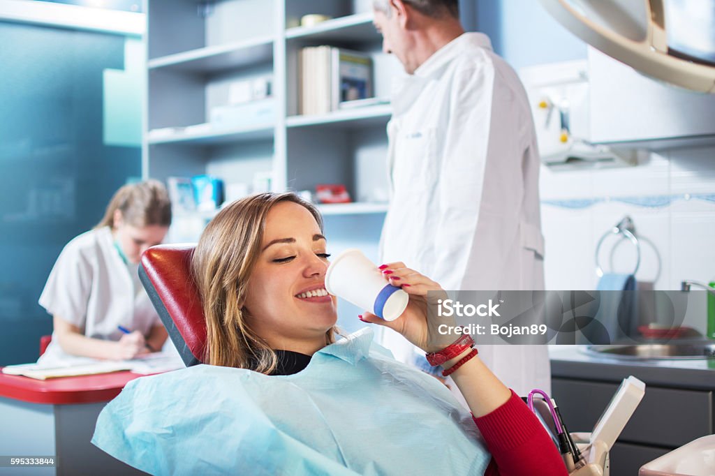 Young woman is taking a glass of water at dentist's. Smiling woman is taking a glass of water in dental office. Dentists in the background. Mouthwash Stock Photo