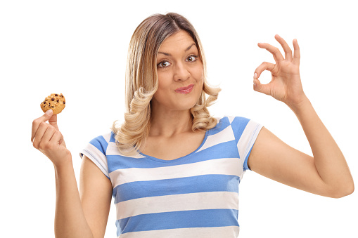 Cheerful woman eating a cookie and making an ok sign isolated on white background