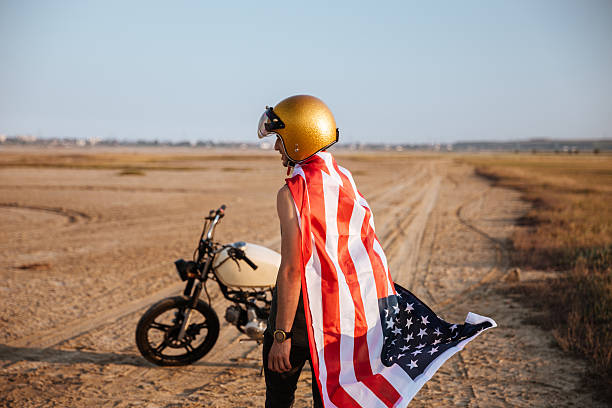 Man wearing american flag cape and golden helmet Young brutal man wearing american flag cape and golden helmet standing near a motorcycle at the desert cafe racer stock pictures, royalty-free photos & images