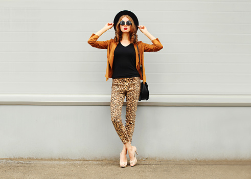 Fashion pretty young woman wearing a retro elegant hat, sunglasses, brown jacket and black handbag posing in city over grey background