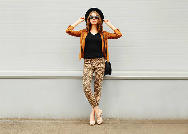 Fashion woman wearing hat, sunglasses, jacket, handbag posing in city Fashion pretty young woman wearing a retro elegant hat, sunglasses, brown jacket and black handbag posing in city over grey background leggings stock pictures, royalty-free photos & images