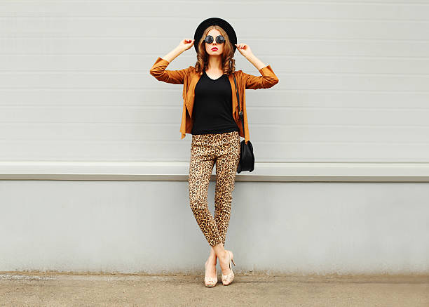 Fashion woman wearing hat, sunglasses, jacket, handbag posing in city Fashion pretty young woman wearing a retro elegant hat, sunglasses, brown jacket and black handbag posing in city over grey background leopard pants outfit stock pictures, royalty-free photos & images