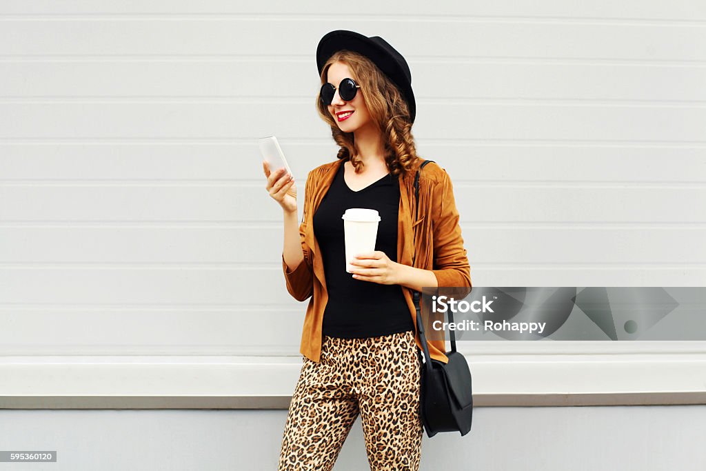 Autumn luxury pretty woman, coffee cup, smartphone walking in city Autumn luxury look, pretty cool smiling young woman with coffee cup using smartphone walking in city, happy female model wearing elegant hat, brown jacket outdoors over grey background Retail Stock Photo